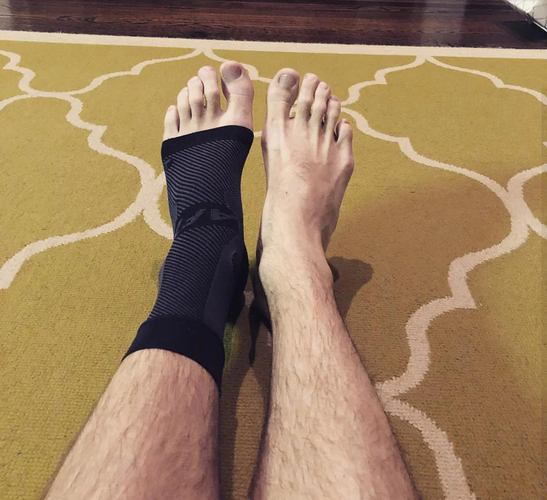 Man's bare legs wearing one OS1st AF7 Ankle Bracing Sleeve in black on the left foot, providing targeted compression and support to relieve ankle pain and instability, designed to improve mobility and enhance performance.