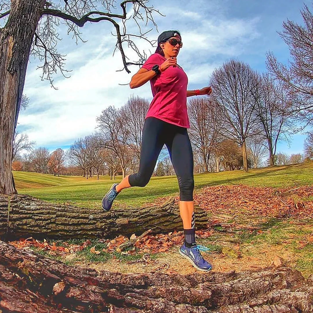 Lady running in grassy area, jumping over log wearing OS1st AF7 Ankle Bracing Sleeve on left foot providing targeted compression and support to relieve ankle pain and instability, designed to improve mobility and enhance performance.