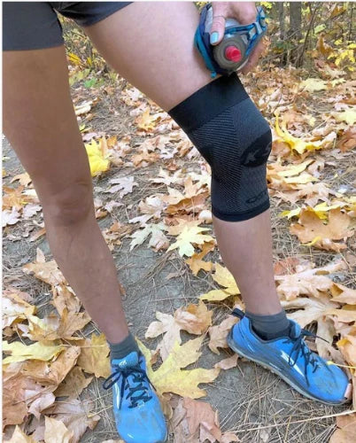 Runner standing wearing a OS1st KS7 Performance Knee Sleeve in black, offering targeted compression to support knee pain relief, improve mobility, and enhance performance during physical activity.
