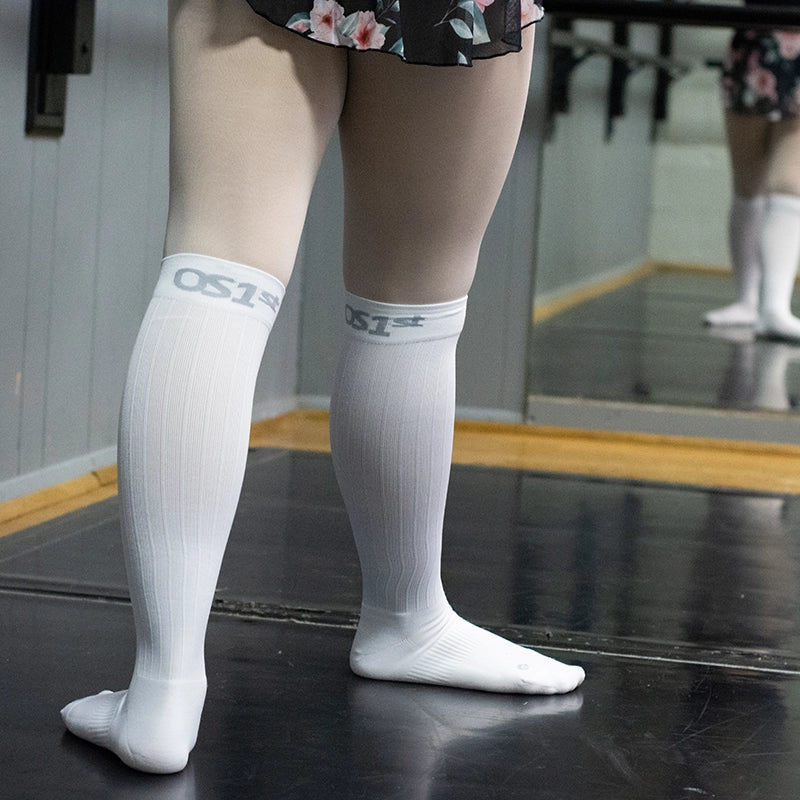 Dancer standing front of bar wearing OS1st TS5 Travel Socks in white, providing compression support to improve circulation and reduce discomfort during travel and long periods of sitting.